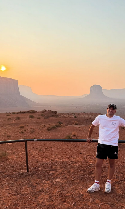 Sam met een wit t-shirt, zwarte korte broek, witte schoenen en zwarte pet staat voor de iconische rotspartijen van Monument Valley. De zon gaat onder.