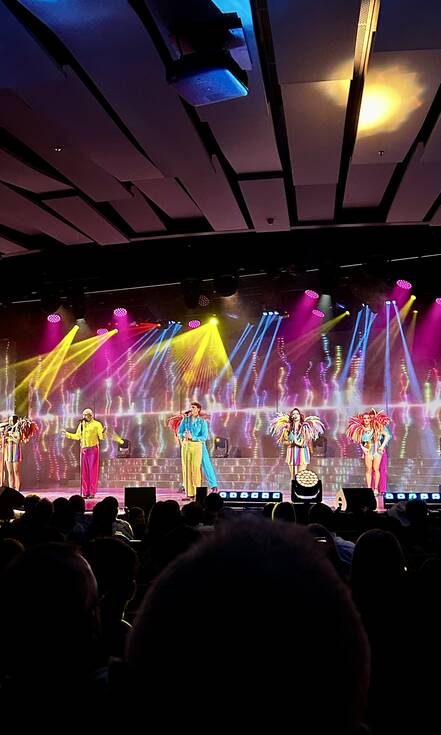 Op de foto zie je een show met blauw, geel en roze lichten. Er staan vijf personen op het podium in kleurrijke outfits. Sommigen zingen in een microfoon.