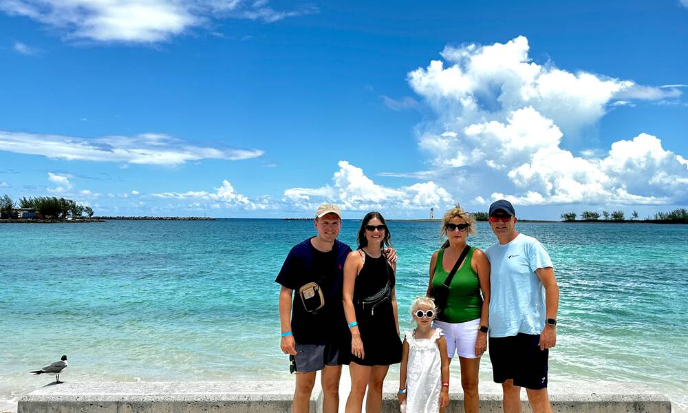 Op de foto zie je Carola en haar familie. Van links naar rechts, de man van Carola met pet, Carola, dochterlief, de moeder van Carola en haar stiefvader ook met pet. De achtergrond is de oceaan met blauw water. 