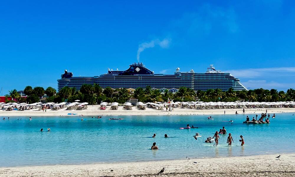 Op de foto zie je een strook blauw water, wit zandstrand met netjes gerangschikte parasols en daarachter een groot cruiseship